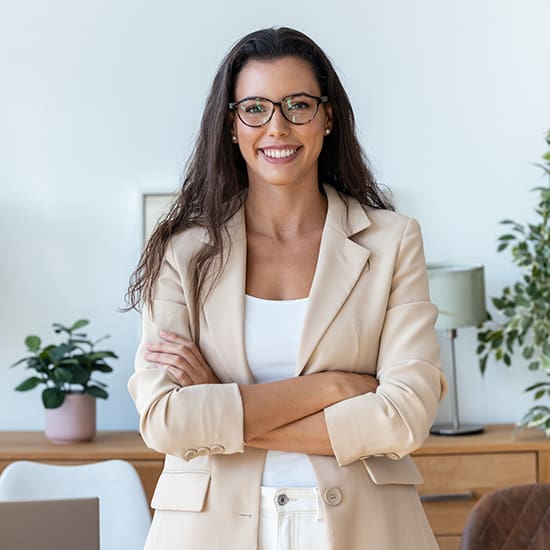 A woman with long dark hair wearing glasses and a beige blazer stands confidently with her arms crossed. She is smiling and standing in a modern, well-lit room with plants and a wooden sideboard in the background.