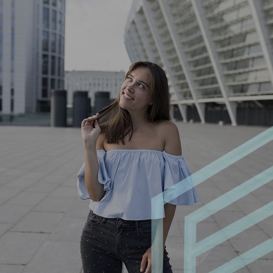 A woman with long brown hair and an off-the-shoulder blue top stands on a large paved area, twirling a strand of her hair and smiling while looking upward. She is in front of a modern, curved glass building on a cloudy day.