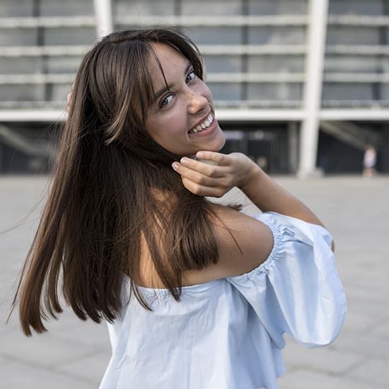 A person with long brown hair, wearing an off-the-shoulder light blue top, is smiling and looking over their shoulder while touching their hair. They are standing outside with a modern building in the background.