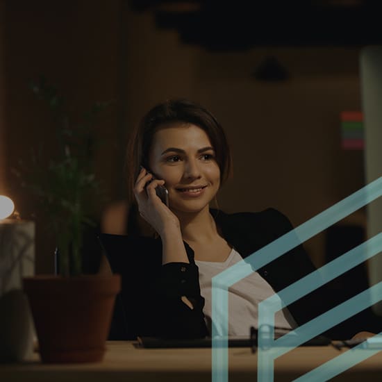 A young woman with brown hair is sitting at a desk, smiling while talking on the phone. She is wearing a black blazer and white top. The room is dimly lit, and there's a plant and a lamp on the desk. A geometric graphic overlay appears in the foreground.
