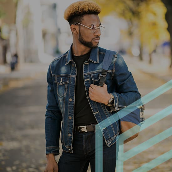 A man with a blonde high-top haircut and glasses, wearing a denim jacket over a black shirt, stands in an outdoor urban setting. He is holding a strap of a backpack on his shoulder and looking to his right. The background features trees with autumn leaves.