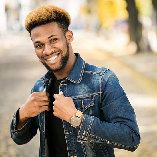 A man stands outdoors on a tree-lined path, smiling at the camera. He has short, styled hair and wears a denim jacket over a black shirt. He holds the jacket's lapels with both hands, and a watch is visible on his left wrist. The background is softly blurred.