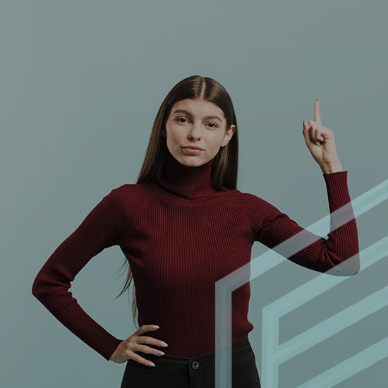 A woman with long brown hair, wearing a red turtleneck and black pants, stands against a gray background. She holds up one hand with her index finger pointing upward while her other hand rests on her hip. She appears confident and focused.
