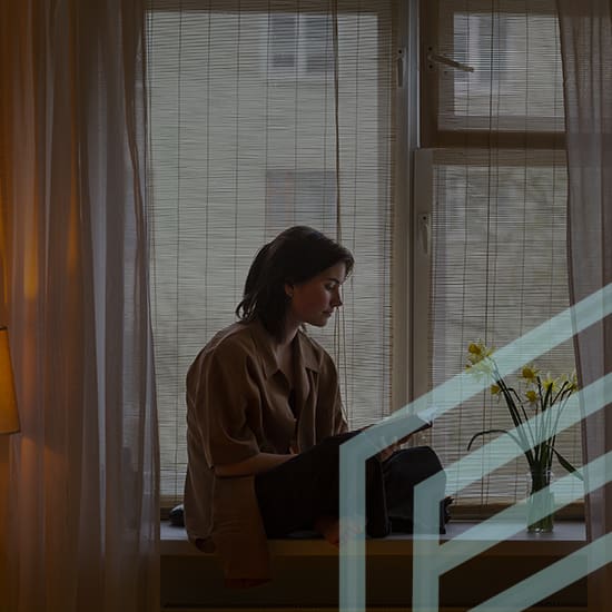 A person with short dark hair sits on a windowsill reading a book, framed by a softly lit room with sheer curtains. A vase with yellow flowers and a lit floor lamp are visible beside them. The window is partially covered with bamboo blinds, and the outdoor scene is blurred.