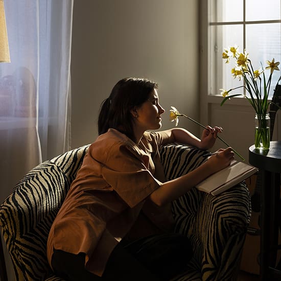 A person sits in an armchair with zebra-striped upholstery, holding a yellow flower while writing in a notebook. Sunlight streams through a nearby window, illuminating them and a vase of yellow flowers on the table beside them.