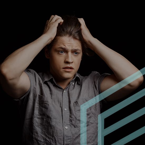 A young man with blue eyes and brown hair, wearing a short-sleeved, button-up shirt, looks distressed as he pulls at his hair with both hands. The background is black with a partially visible geometric design in light blue in the bottom right corner.