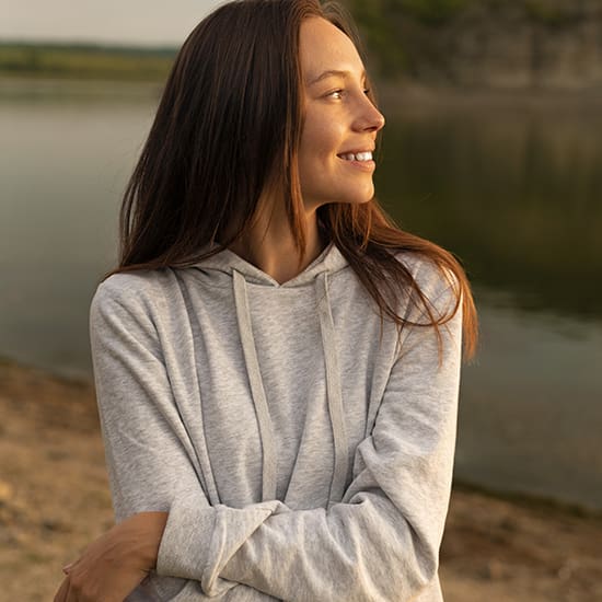 A young woman with long brown hair smiles while looking to her right. She is standing outdoors near a calm body of water, wearing a light grey hoodie. The background includes a shoreline with greenery and cliffs in the distance.