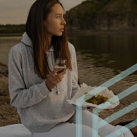 A young woman in a gray hoodie sits on a blanket by the water, holding a glass of white wine in one hand and a plate of grapes in the other. She looks off into the distance with a serene expression, and the background features a rocky shoreline and greenery.