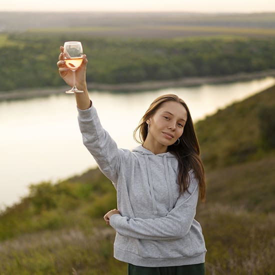 A woman with long hair, wearing a gray hooded sweatshirt, stands outdoors by a river at sunset. She is smiling and holding up a glass of rosé wine in celebration. The background features lush green hills and a calm river.
