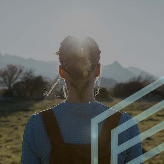 A person with light brown hair, wearing a blue top and brown overalls, stands in a field facing away, with the sun shining over their head. Mountains are visible in the background. The image features a semi-transparent geometric design overlay.