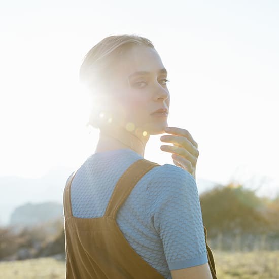 A person with light hair stands outdoors, illuminated by the sun shining from behind. They are looking over their shoulder, hand gently touching their face. They wear a blue textured shirt and brown overalls, with a hazy landscape visible in the background.