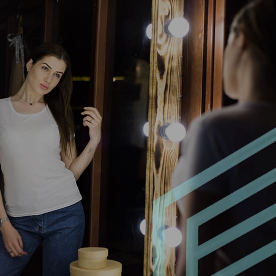 A woman wearing a white t-shirt and blue jeans is standing in front of an illuminated mirror, looking at her reflection. The mirror has round light bulbs along its wooden frame. The background is dimly lit.