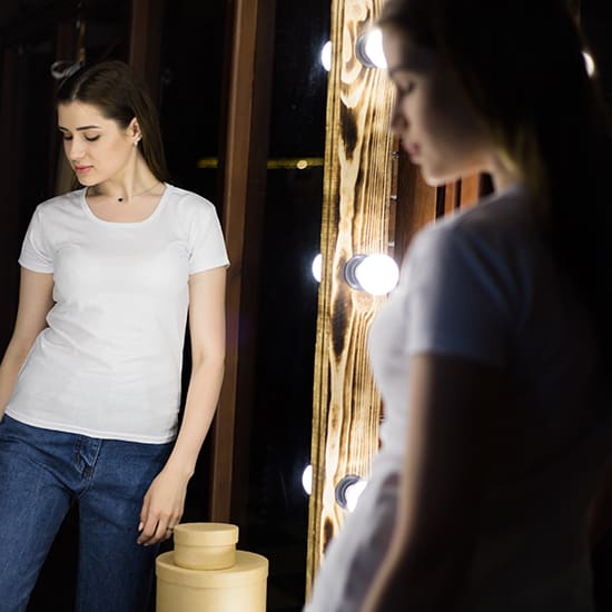 A woman wearing a white t-shirt and blue jeans stands in front of a mirror adorned with lights, looking at her reflection. She has long, straight hair and her expression appears contemplative. The wooden frame of the mirror contrasts with the dark background.