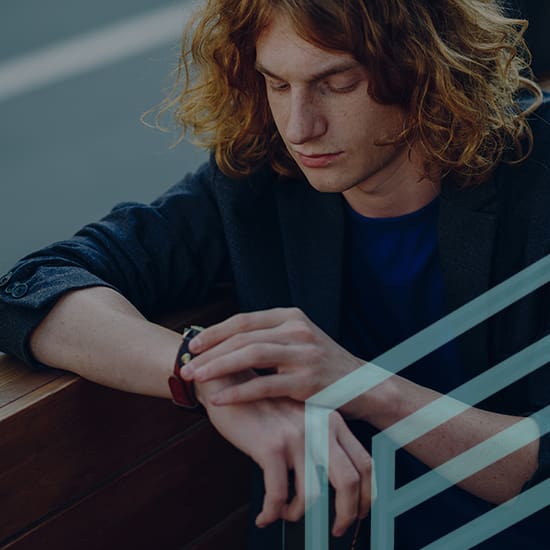 A person with long, wavy, reddish hair, wearing a dark blazer and blue shirt, is sitting and looking at their smartwatch. They have one arm propped up on a wooden bench, with a glass railing in the foreground.
