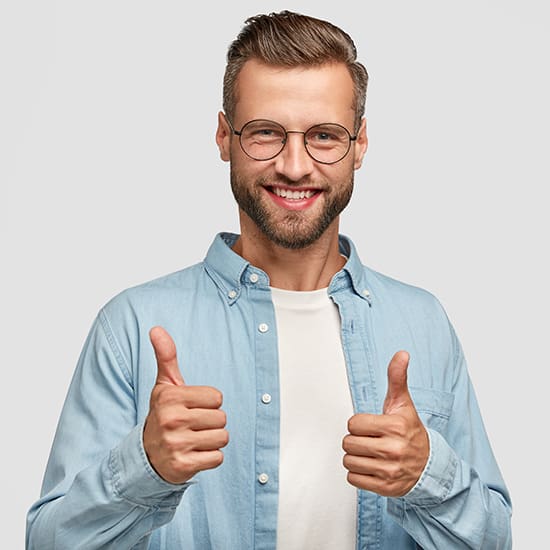A man with a beard and glasses wearing a light blue shirt over a white t-shirt smiles and gives a double thumbs-up gesture. He stands against a plain, light gray background.