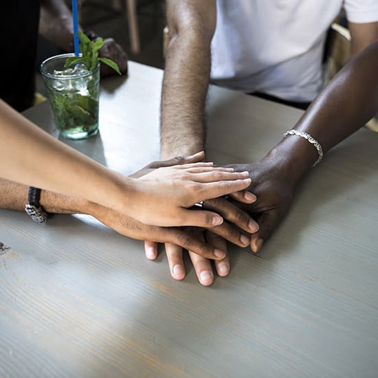 A close-up of five hands of diverse skin tones stacked on top of each other on a wooden table, symbolizing unity and teamwork. In the background, a glass of green beverage with a straw is partially visible.