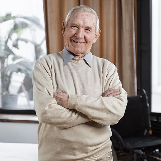An elderly man with short white hair smiles at the camera, standing with his arms crossed. He is wearing a beige sweater over a collared shirt. In the background, there are large windows with light curtains, and a wheelchair is partially visible.