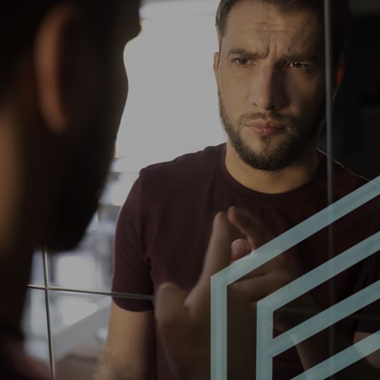 A man with a beard wearing a maroon shirt touches a geometric pattern on a mirror while looking at his reflection thoughtfully. The background is softly lit, with indistinct items visible.