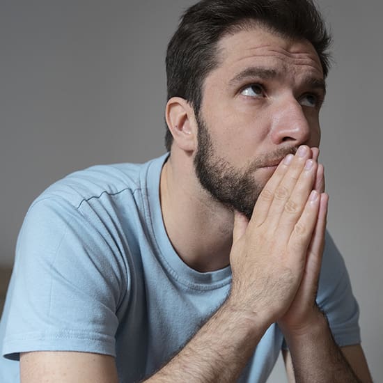 A man with a beard and short hair sits with his hands clasped near his face, appearing deep in thought. He is wearing a light blue T-shirt and looking slightly upward with a serious expression against a plain background.