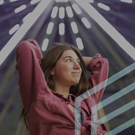 Image of a woman with long brown hair looking up with a thoughtful expression. She has her arms raised and hands behind her head. She is wearing a red shirt and standing against a background of bright, radiating lights.