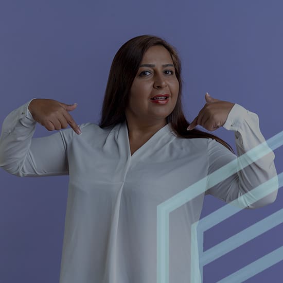 A woman with long dark hair stands against a purple background, wearing a white blouse. She is pointing to herself with both hands, suggesting confidence or self-identification. The image has diagonal white stripes in the bottom right corner.