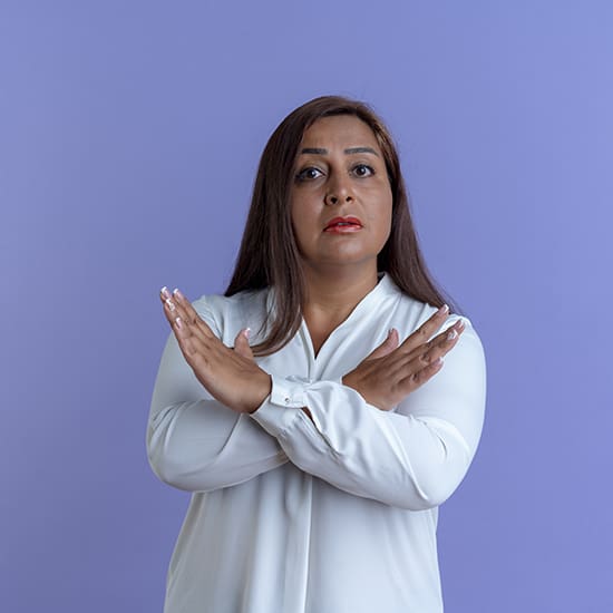 A woman with long brown hair wearing a white blouse stands against a purple background. She has a serious expression and is crossing her arms in front of her chest, forming an 'X' with her hands.