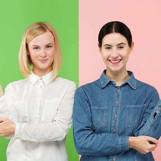 Two women stand side by side. The woman on the left has blonde hair and wears a white shirt against a green background. The woman on the right has dark hair and wears a denim shirt against a pink background. Both women are smiling.