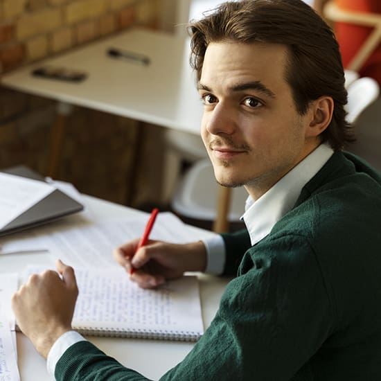 A young man with shoulder-length brown hair, wearing a green sweater over a white shirt, sits at a desk. He is holding a red pen and writing in a notebook, looking up with a slight smile. The background shows a blurred interior with a table and a brick wall.