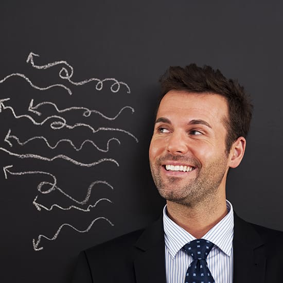 A smiling man in a suit and tie stands in front of a chalkboard with several chalk-drawn arrows and squiggles pointing in different directions. The man appears to be looking at the drawings with amusement or interest.