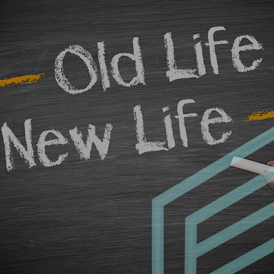 A hand holding a piece of chalk is drawing on a blackboard. The words "Old Life" and "New Life" are written in white chalk above and below a dividing line. The concept suggests starting anew or transitioning from an old phase to a new one.
