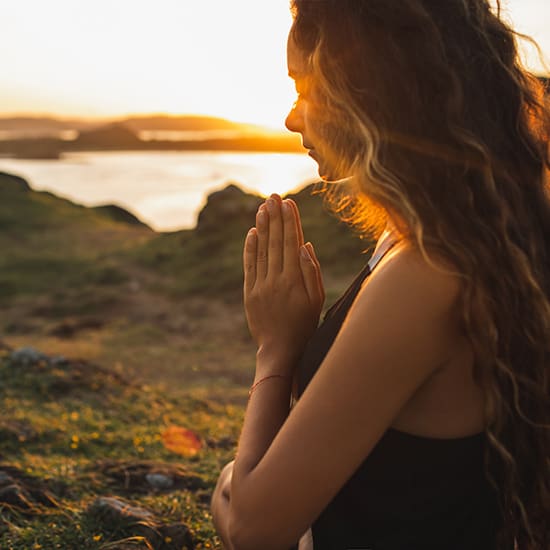 A person with long, wavy hair stands outdoors at sunset, hands in a prayer position close to their chest. They are facing to the right, with the serene landscape and a body of water in the background. The sunlight creates a warm glow around them.