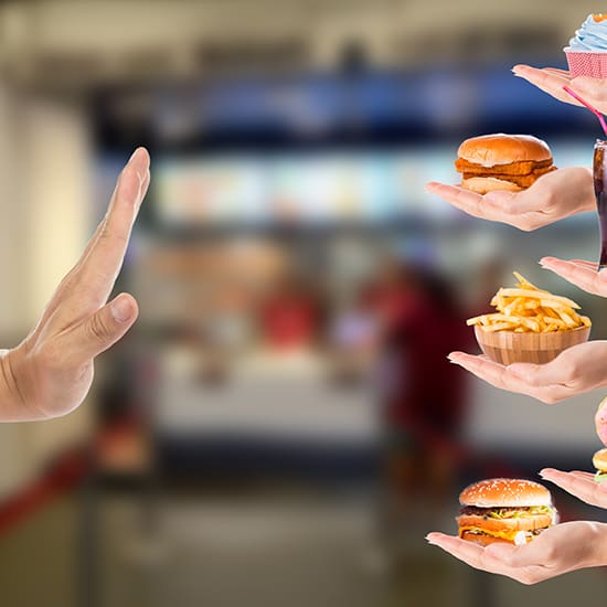 A person's hand is shown in a "stop" gesture in the foreground, facing several hands holding an assortment of fast food items, including burgers, fries, and a soda. The background is blurry, resembling a fast-food restaurant.