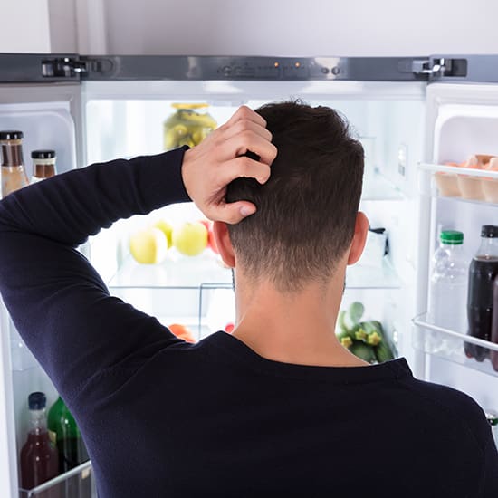 A man stands with his back to the camera, scratching his head while looking into an open refrigerator. The fridge shelves are stocked with various items, including bottles and fresh produce. The scene suggests he might be deciding what to eat.