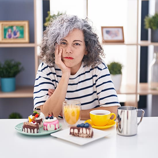A woman with curly gray hair sits at a table looking pensive. She is wearing a striped shirt and resting her face on her hand. On the table, there are various desserts, a glass of juice, a cup of tea or coffee, and a small jug. Shelves and plants are in the background.
