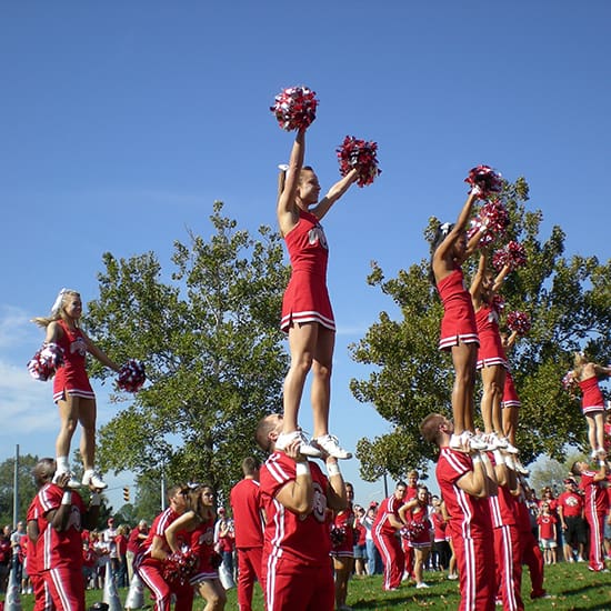 A group of cheerleaders in red uniforms perform a stunt outdoors. Each of the four cheerleaders is lifted by two bases, raising pom-poms in the air against a backdrop of green trees and a clear blue sky. Spectators and more cheerleaders are visible in the background.