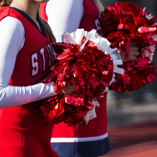 Two cheerleaders in red and white uniforms hold red pom-poms. Only the upper body of the cheerleaders is visible. The background is blurred, focusing attention on the cheerleaders and their pom-poms.