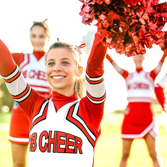 A cheerful cheerleader in a red and white uniform with the text "BB CHEER" on her top, smiles with pom-poms raised in the air. Two other cheerleaders in similar uniforms are in the background, also holding pom-poms. They are outdoors on a sunny day.