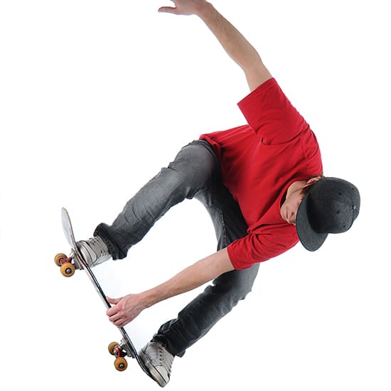 A skateboarder dressed in a red T-shirt, grey pants, and a dark cap performs an aerial trick on a skateboard against a white background. He is grabbing the side of the skateboard with one hand while airborne.