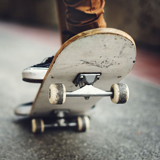 Close-up of a skateboarder in action, with focus on the underside of the skateboard. Only the legs and feet wearing sneakers are visible, along with the worn wheels and deck of the skateboard in motion against a concrete surface.