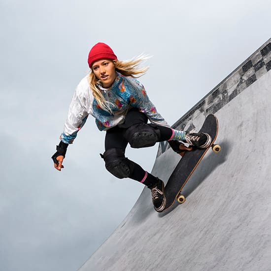 A person in a red beanie, colorful jacket, and knee pads skates on a ramp. The individual is mid-movement, bent low on the skateboard, with hair blowing in the wind against a gray sky backdrop.