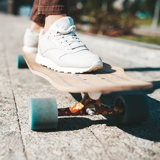Close-up of a person wearing white sneakers, standing on a longboard skateboard outside on a sunny day. The pavement is textured, and greenery is visible in the background, suggesting a park or urban setting.