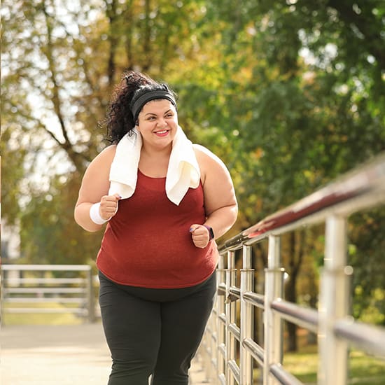 A woman with a towel draped over her shoulders is jogging outdoors on a pathway surrounded by trees. She is wearing a red tank top, black leggings, and a headband, smiling as she exercises. The background shows a metal railing and a sunlit park area.
