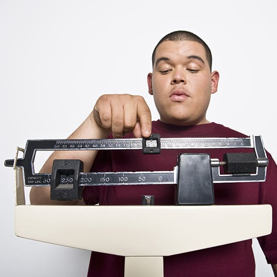 A person in a maroon shirt adjusts the weights on a mechanical scale, looking down at the measurements with a focused expression. The background is plain white.
