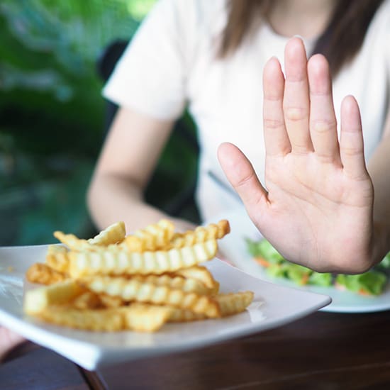 A woman, out of focus in the background, is holding up her hand to refuse a plate of crinkle-cut French fries being offered to her. She is seated at a table with a meal, as blurred green foliage is seen in the distance behind her.