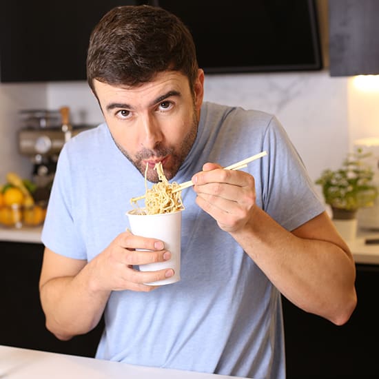 A man with short dark hair, wearing a light blue shirt, is eating noodles from a white takeout container using chopsticks. He is in a modern kitchen with various cooking items and fruits visible in the background.