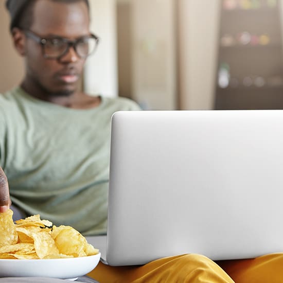 A person wearing glasses and a beanie is sitting and using a laptop on their lap. They are reaching for a potato chip from a bowl in their other hand. The background is slightly blurred, focusing on the person and the laptop.