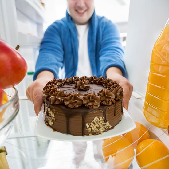 A person wearing a blue robe and white shirt is seen through the open fridge door, smiling and holding a chocolate cake with decorative frosting and nuts on the side. The fridge contains fruits and a bottle of orange juice.