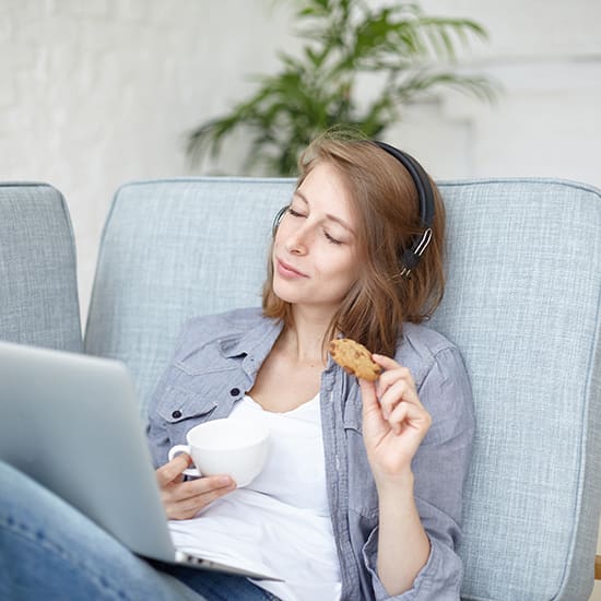 A woman with long hair sits on a light blue couch wearing headphones, holding a cookie in one hand and a white coffee cup in the other. She has a laptop on her lap and appears relaxed. A green plant is visible in the background.
